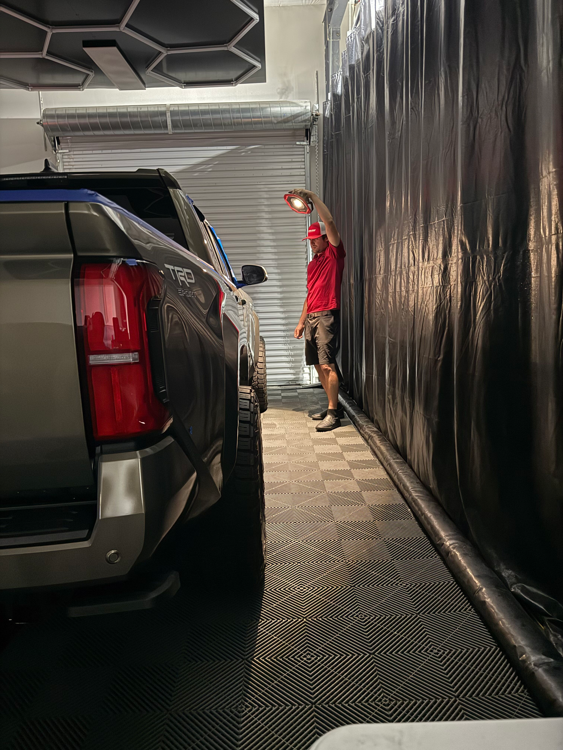 A person in a red shirt washes a dark SUV in a car wash. The person holds a light near the vehicle.