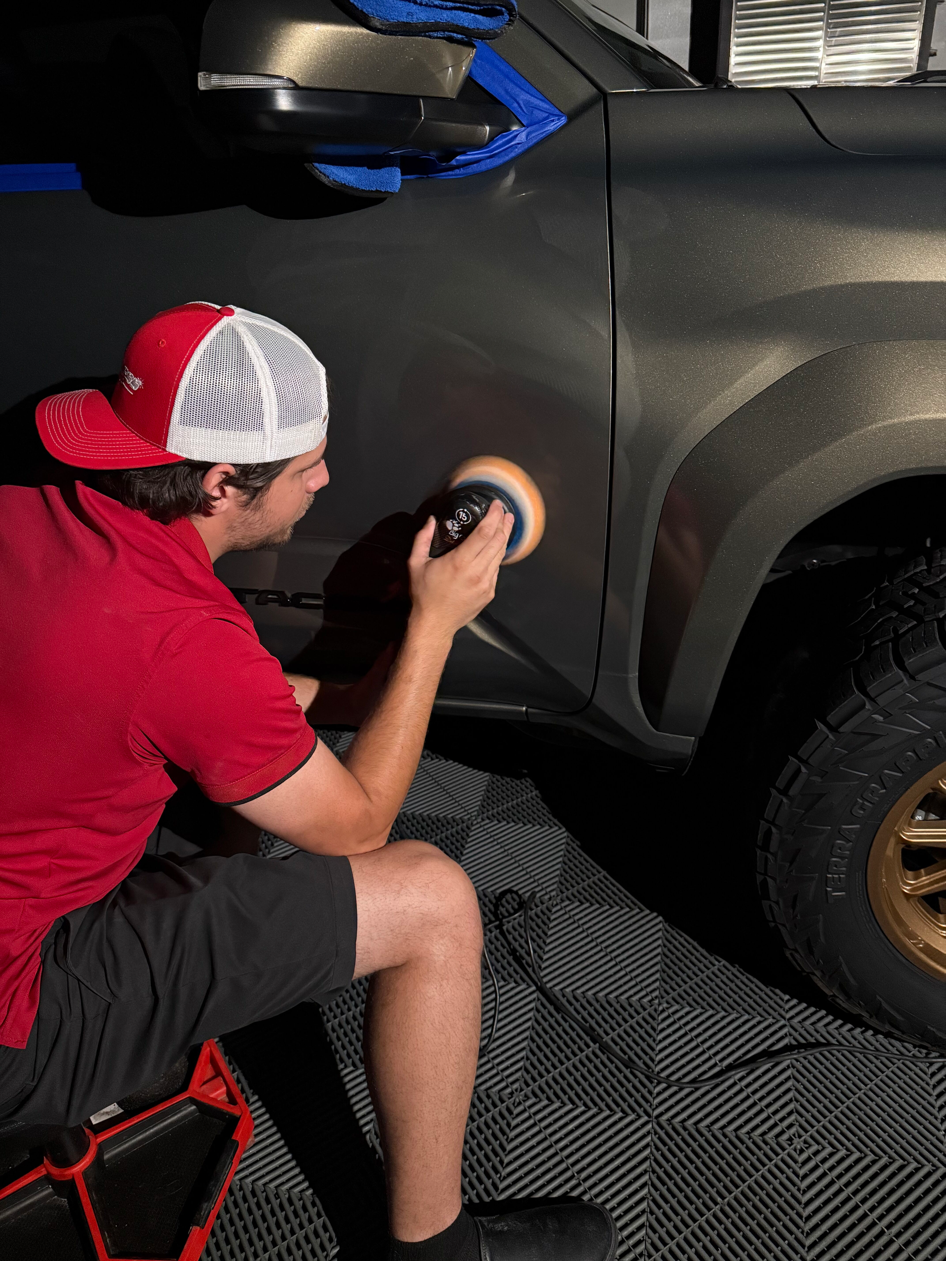 A person polishes a dark gray truck with a power buffer. They wear a red shirt, shorts, and a red and white hat.