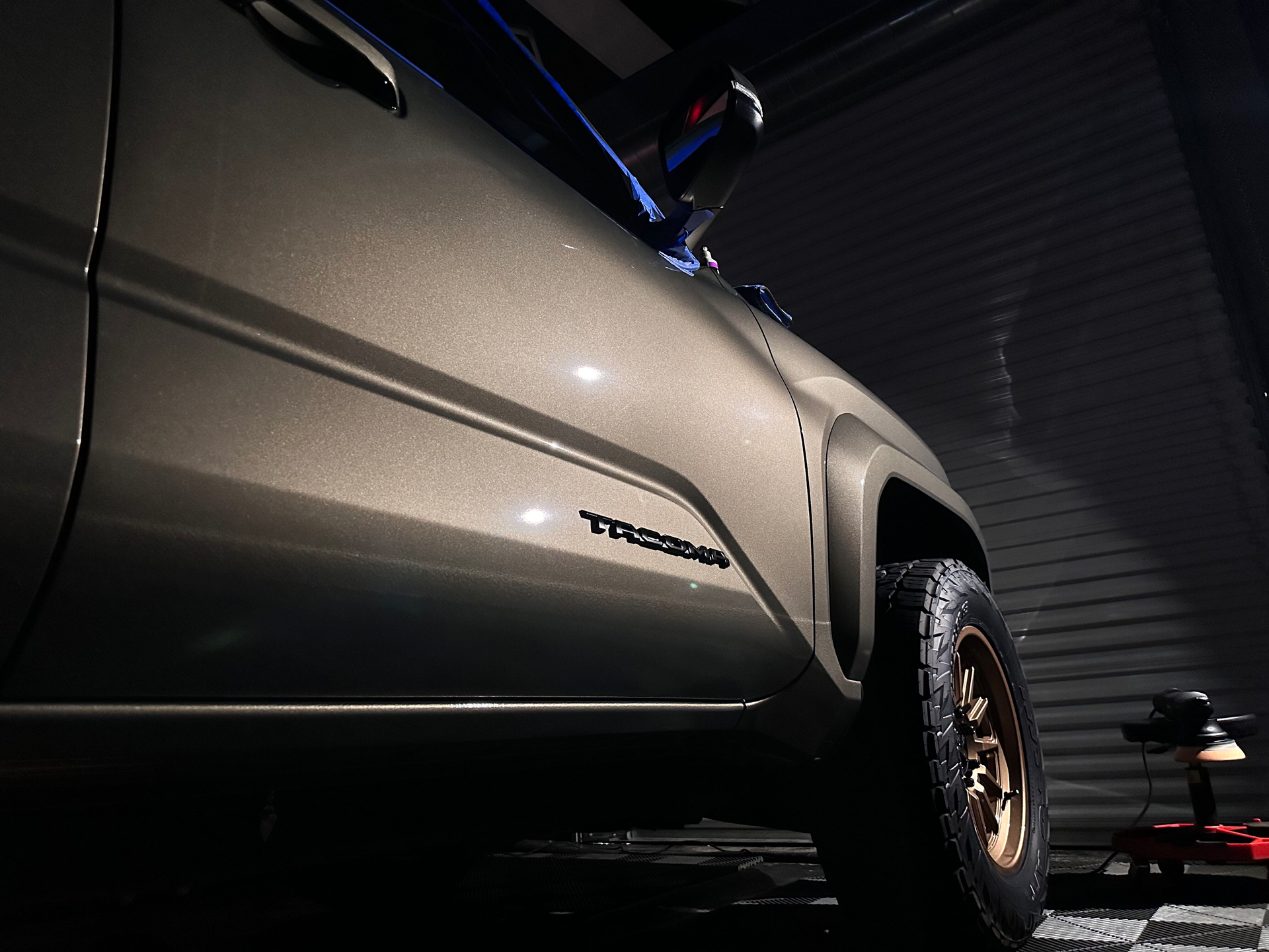 Close-up of a tan Toyota Tacoma truck, with a bronze wheel and tire, in a garage.