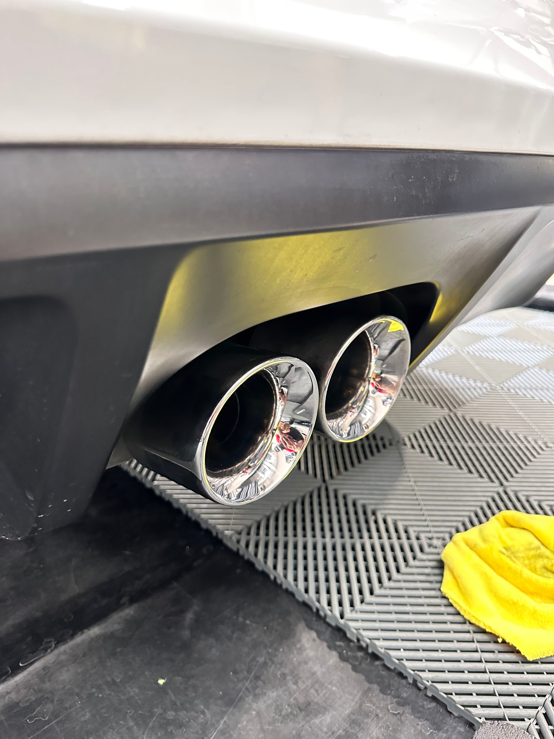 Close-up of a car's dual exhaust pipes. Chrome tips emerge from a black bumper, reflecting light.