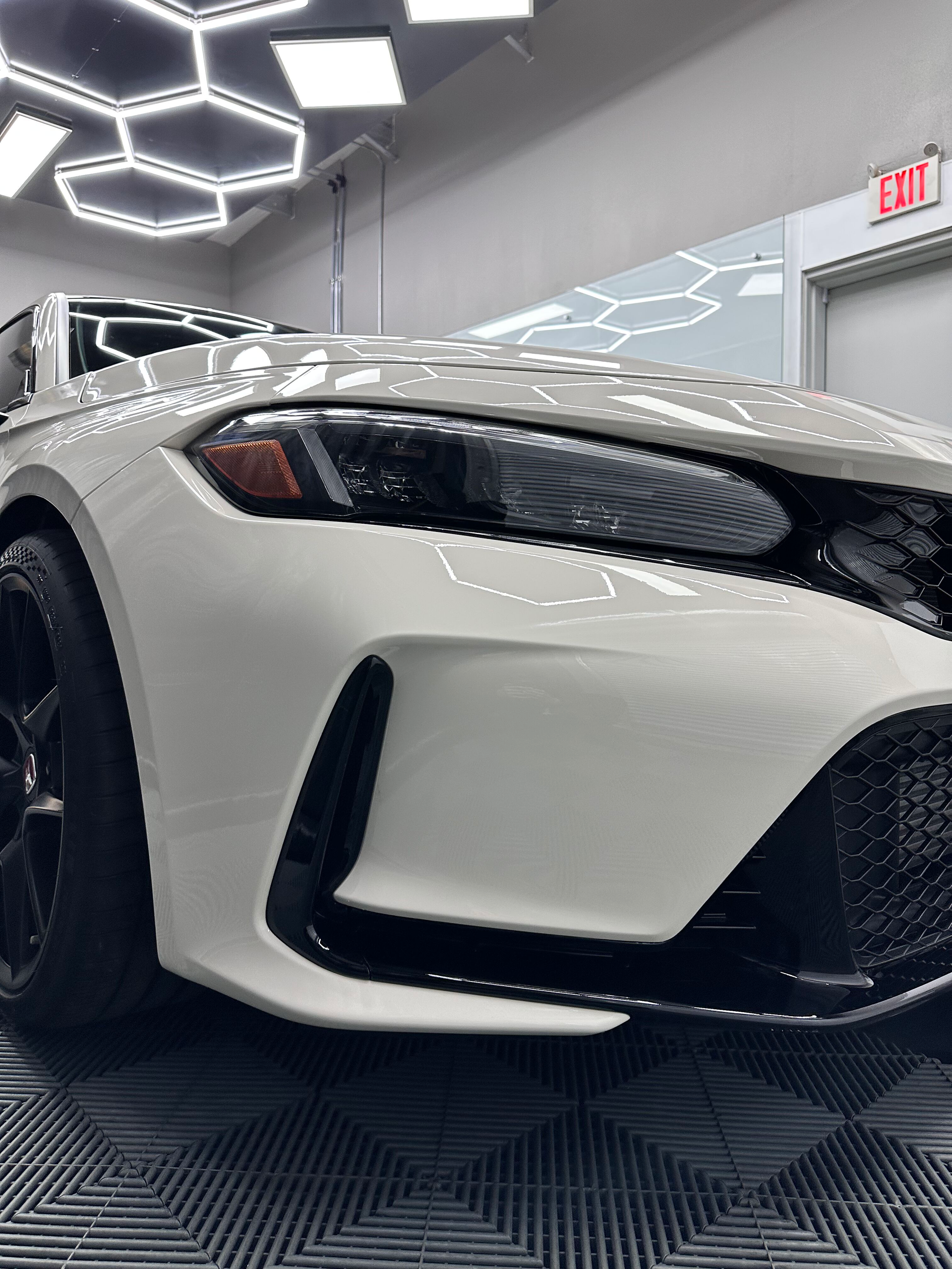 White car in a garage with black accents and black rims. Overhead, honeycomb-shaped lights.