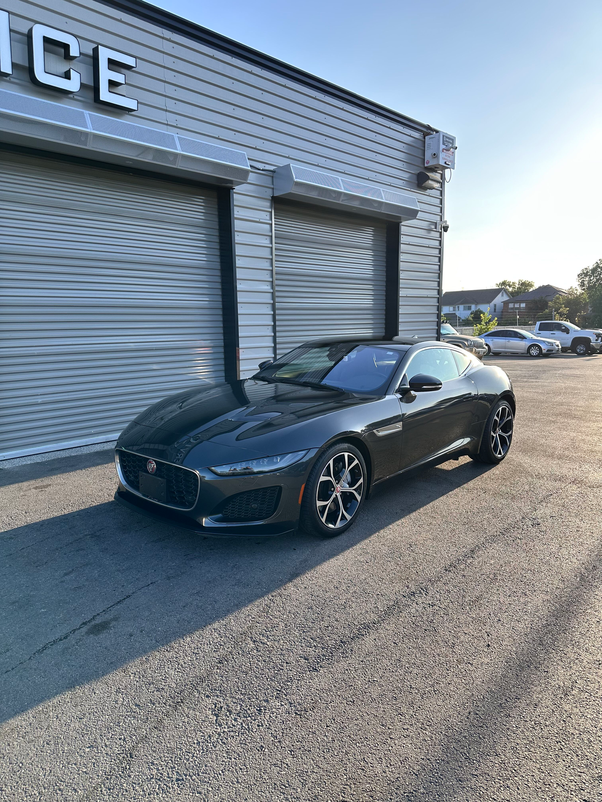 Dark gray Jaguar sports car parked in front of a garage with a closed roll-up door. Sunlight shines.