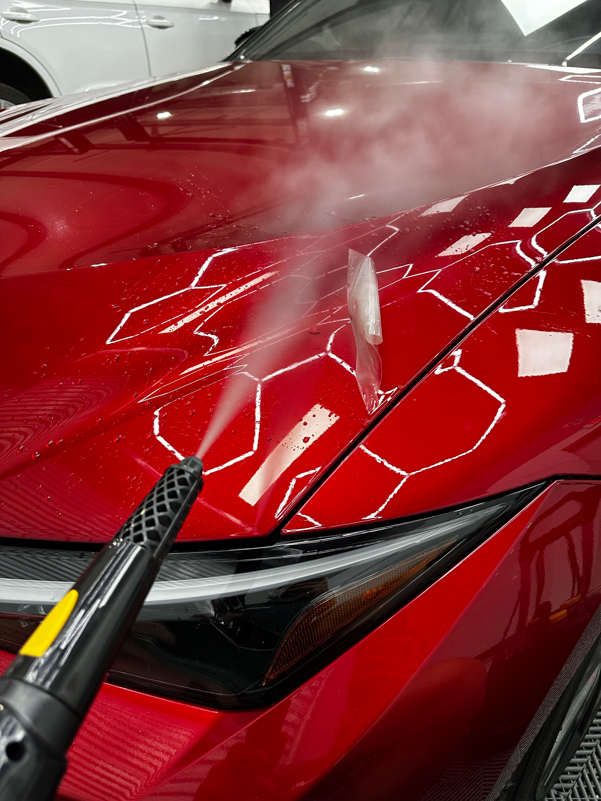 Steam cleaning a red car's hood, highlighting the contours of a protective film with white tape.