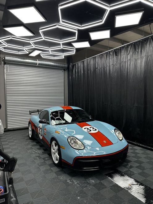 Light blue Porsche Cayman with red racing stripes inside a car detailing bay, under hexagon lights.