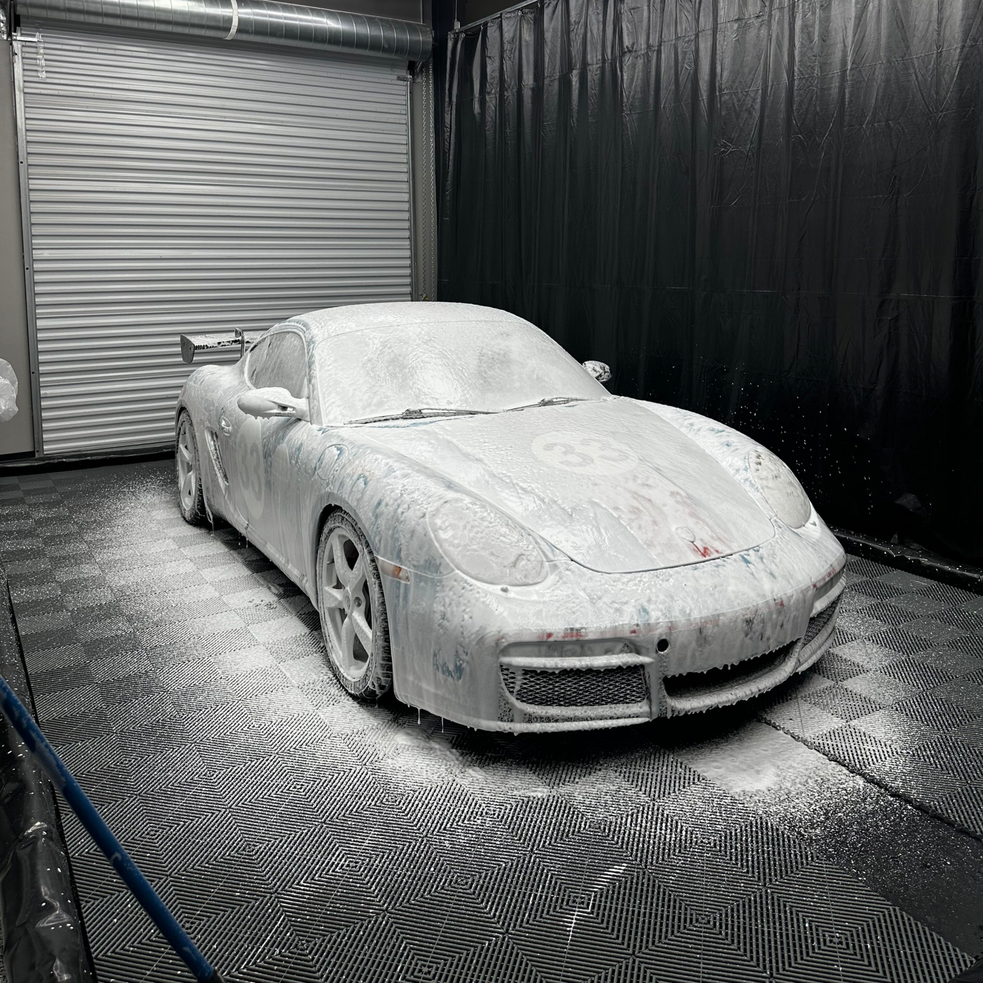 A white sports car covered in white foam inside a car wash bay with hexagon-shaped ceiling lights.