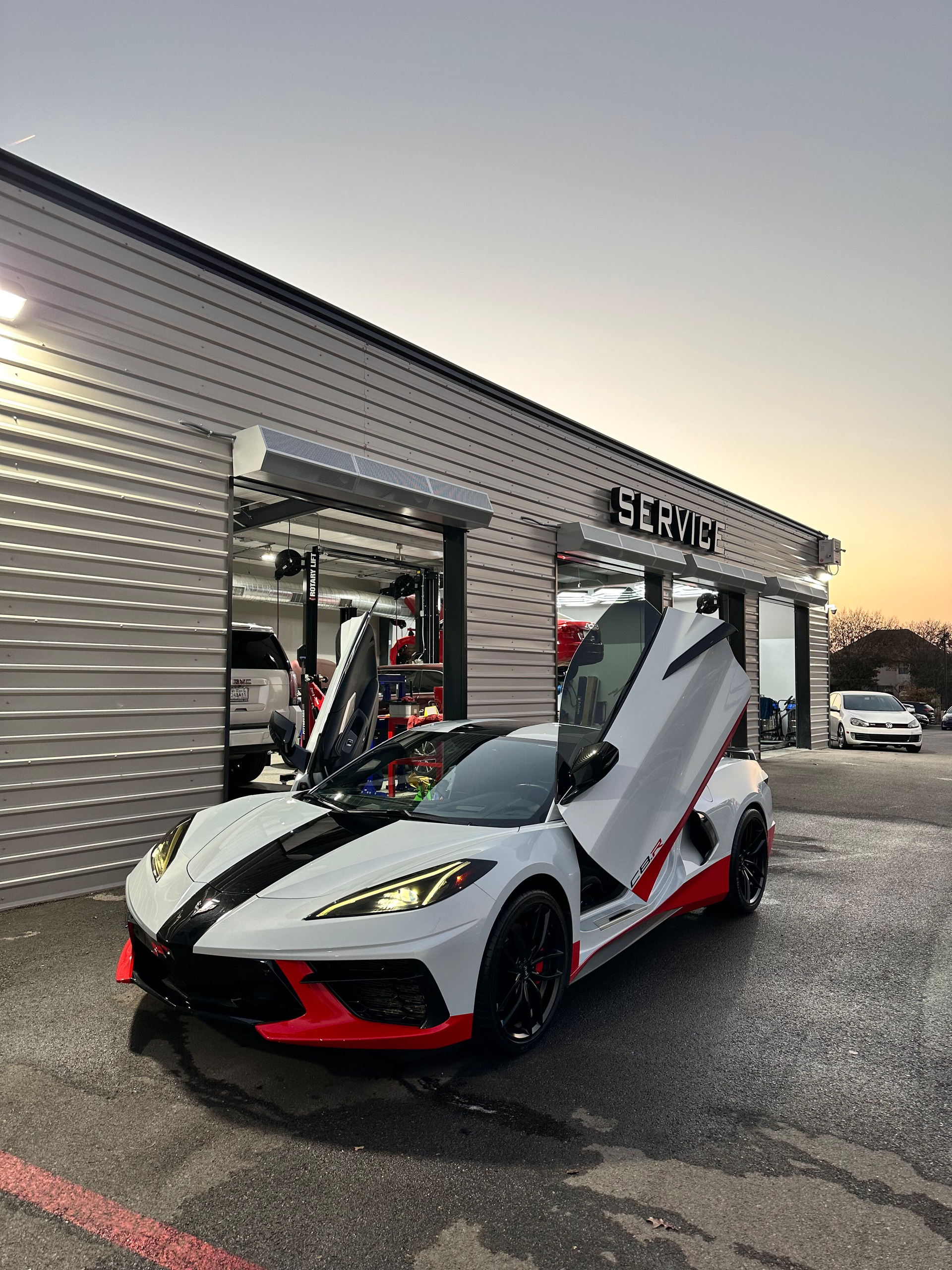 White and black sports car with red accents, doors open, parked in front of a car garage. Evening light.