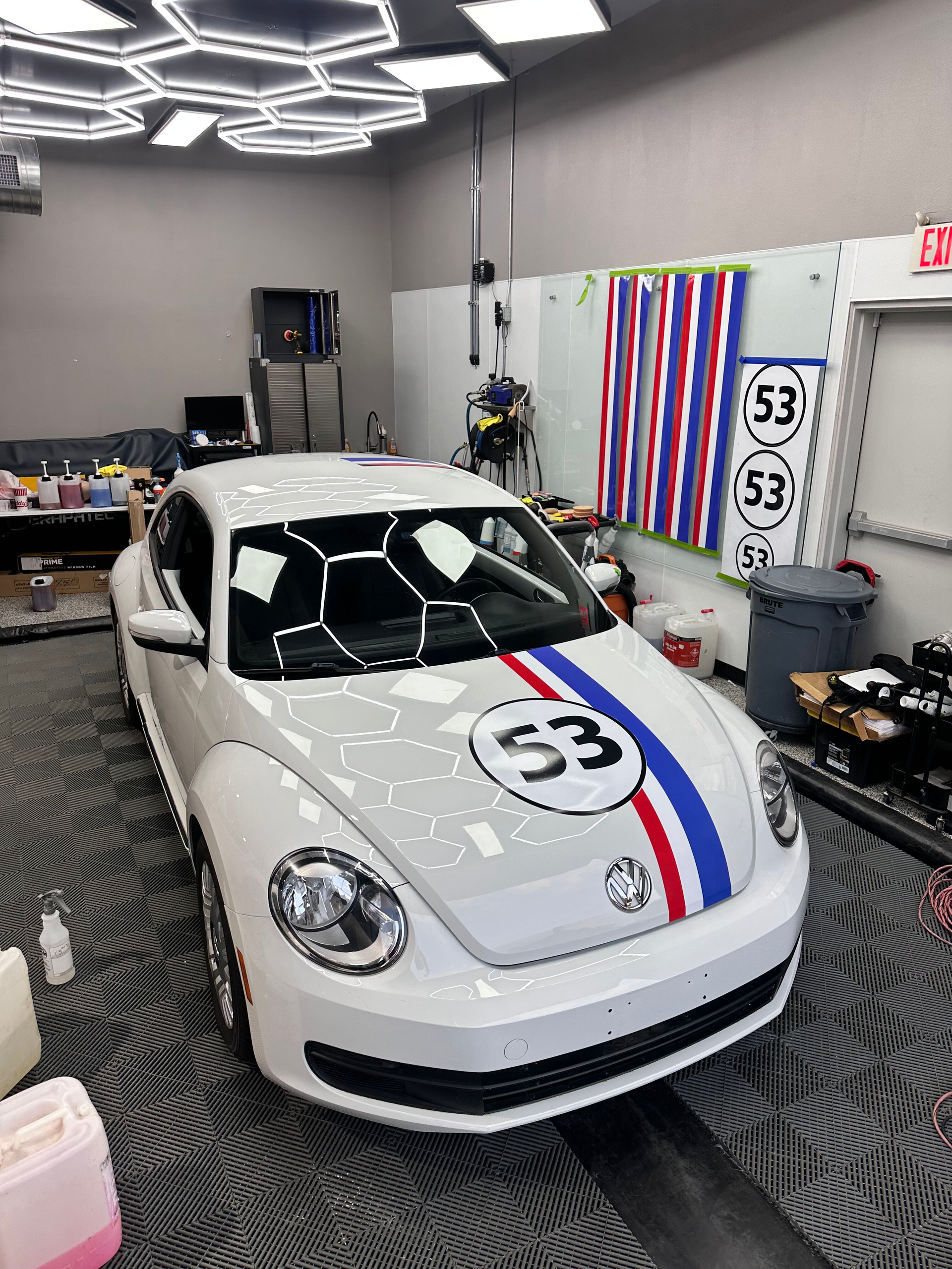 White Volkswagen Beetle with racing stripes and the number 53 in a garage, lit by hexagonal ceiling lights.
