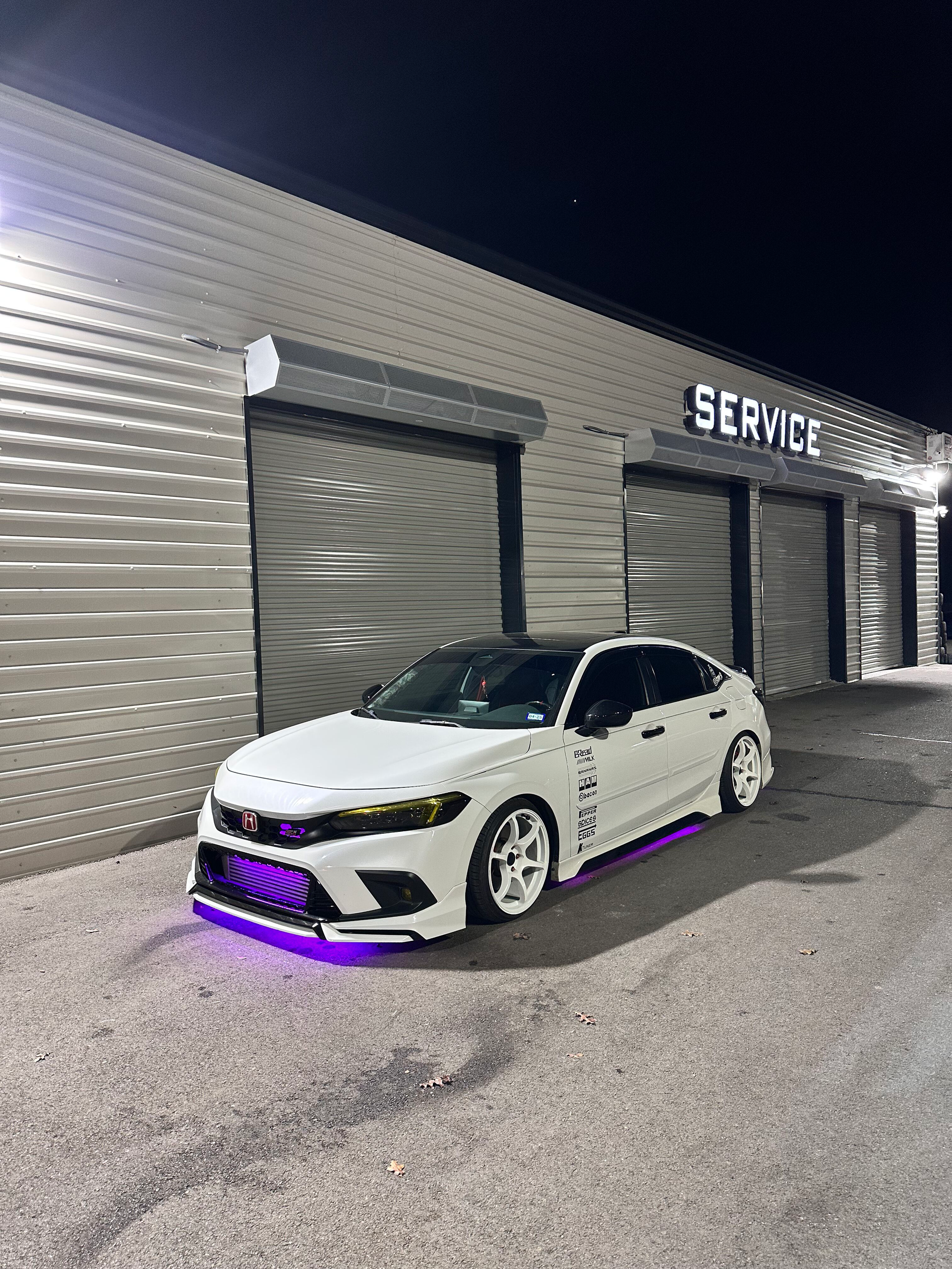 White Honda Civic with purple underglow parked in front of a service garage at night.