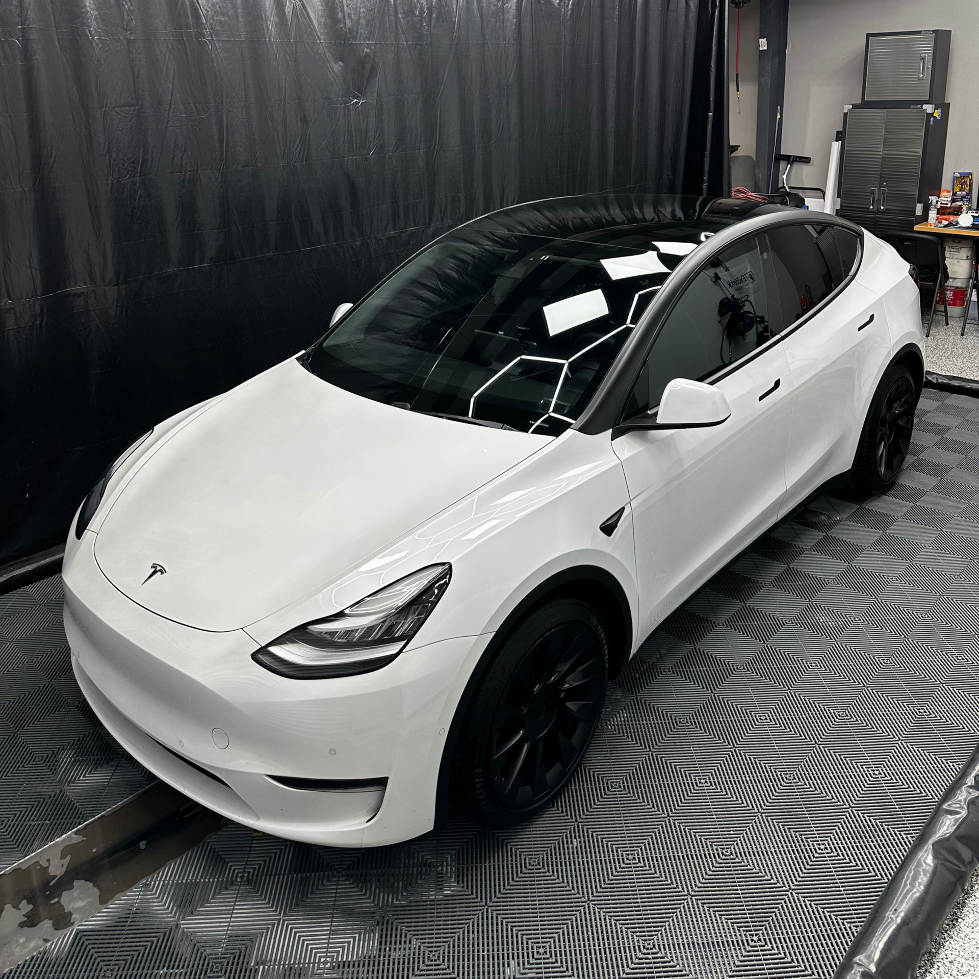 White Tesla Model Y in a garage, illuminated by hexagon lights. The car has a black roof and dark wheels.