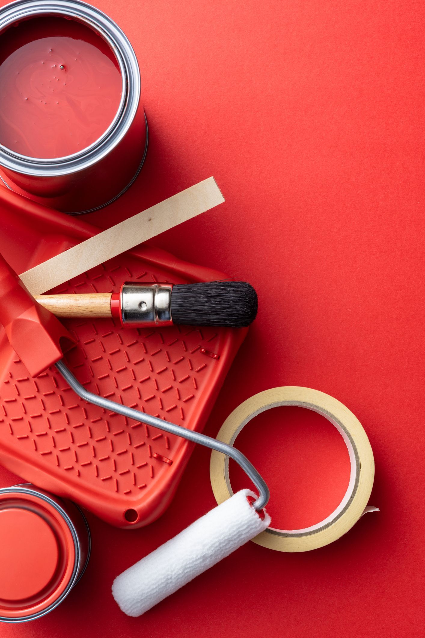 An overhead view of high-quality painting tools available at our Orillia store: an open can of red paint, a paint roller in a tray, a brush, and tape.