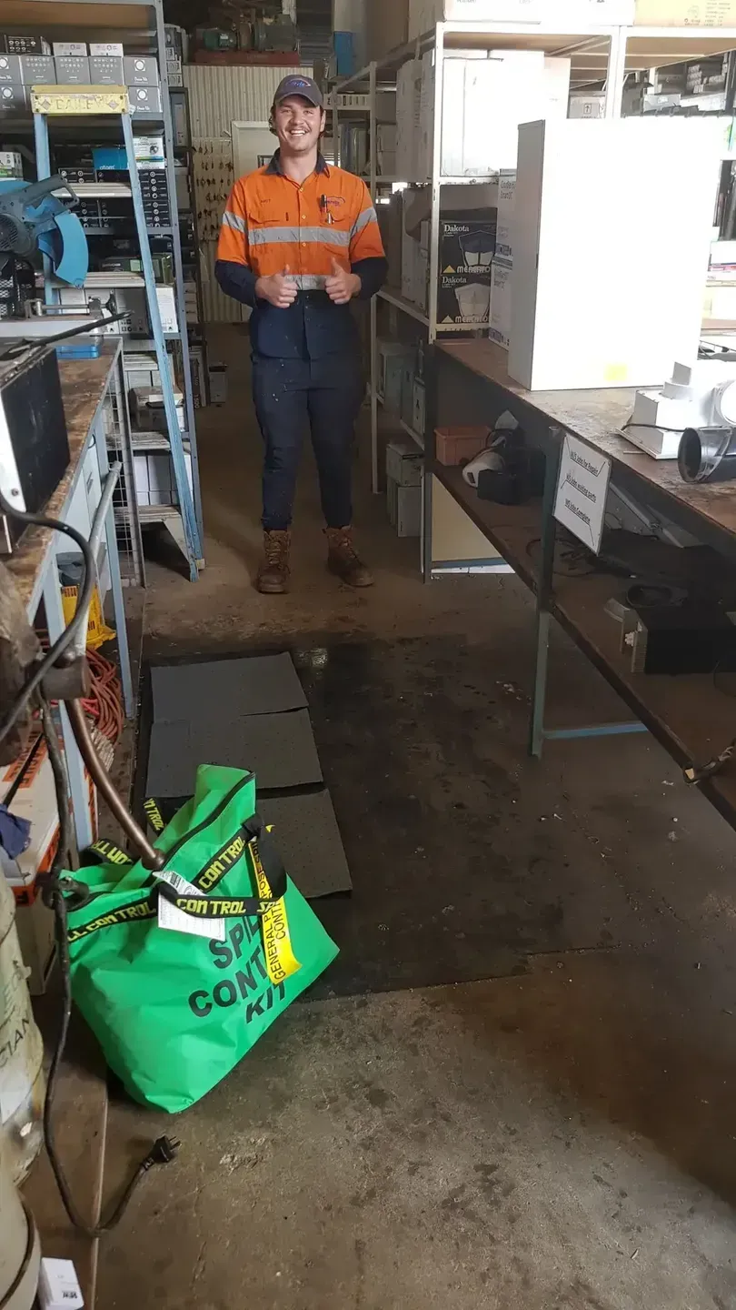 Man in work clothes stands in a workshop, giving a thumbs up, with shelves and tools in the background.