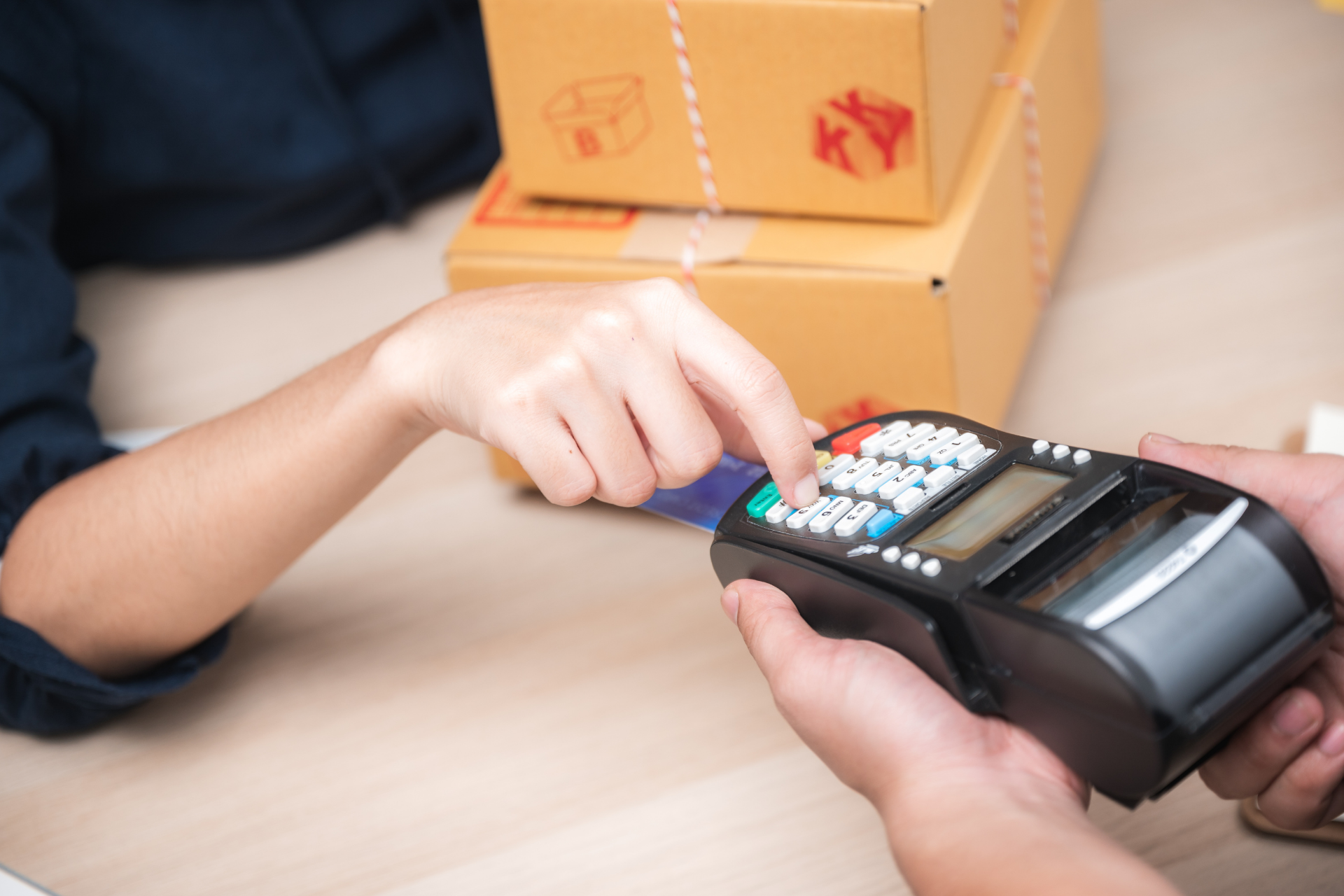 A person uses their finger to enter information into a hand-held card payment terminal near stacked cardboard shipping boxes.