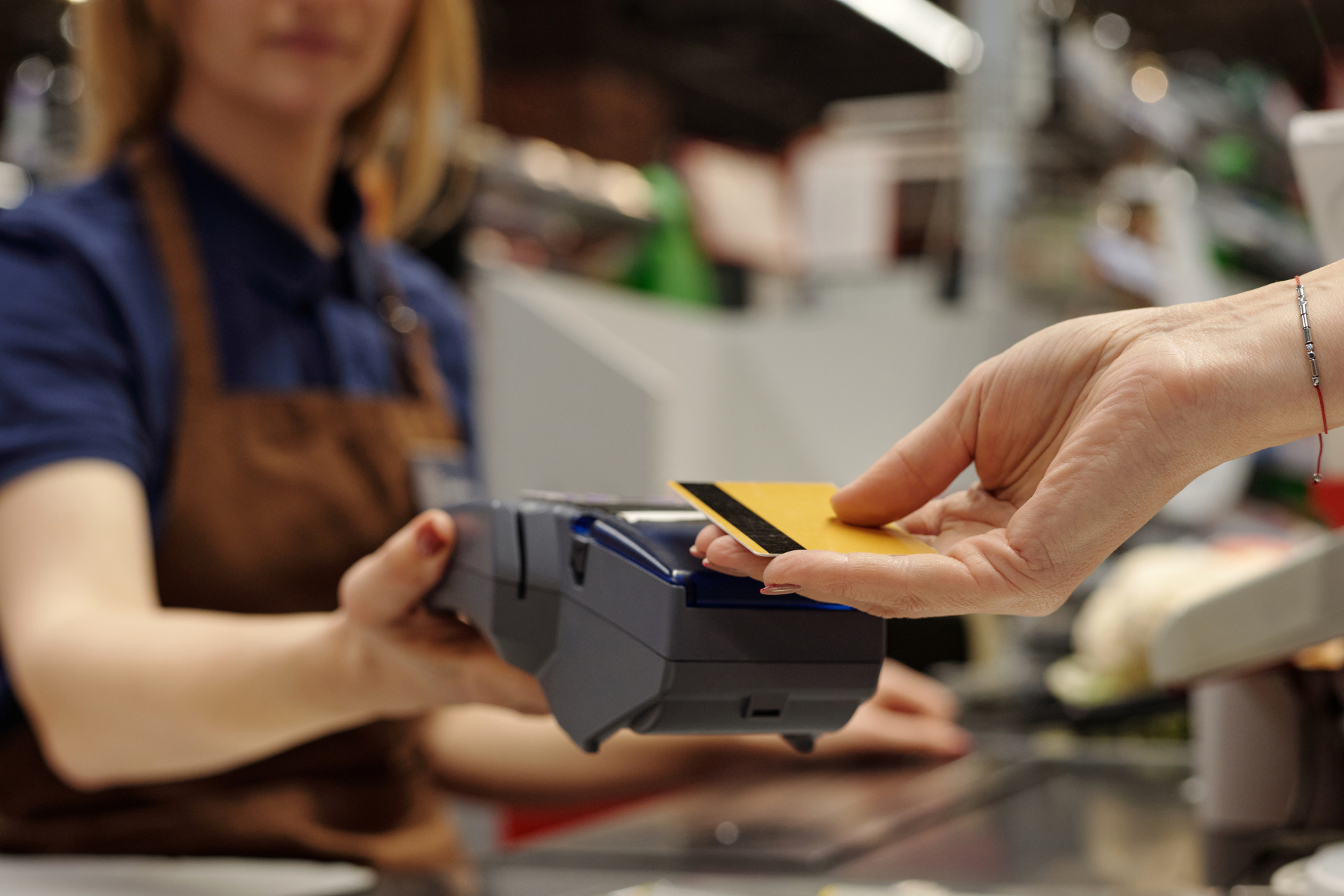 A customer holds a yellow bank card near a card payment terminal held by a retail worker in a brown apron.