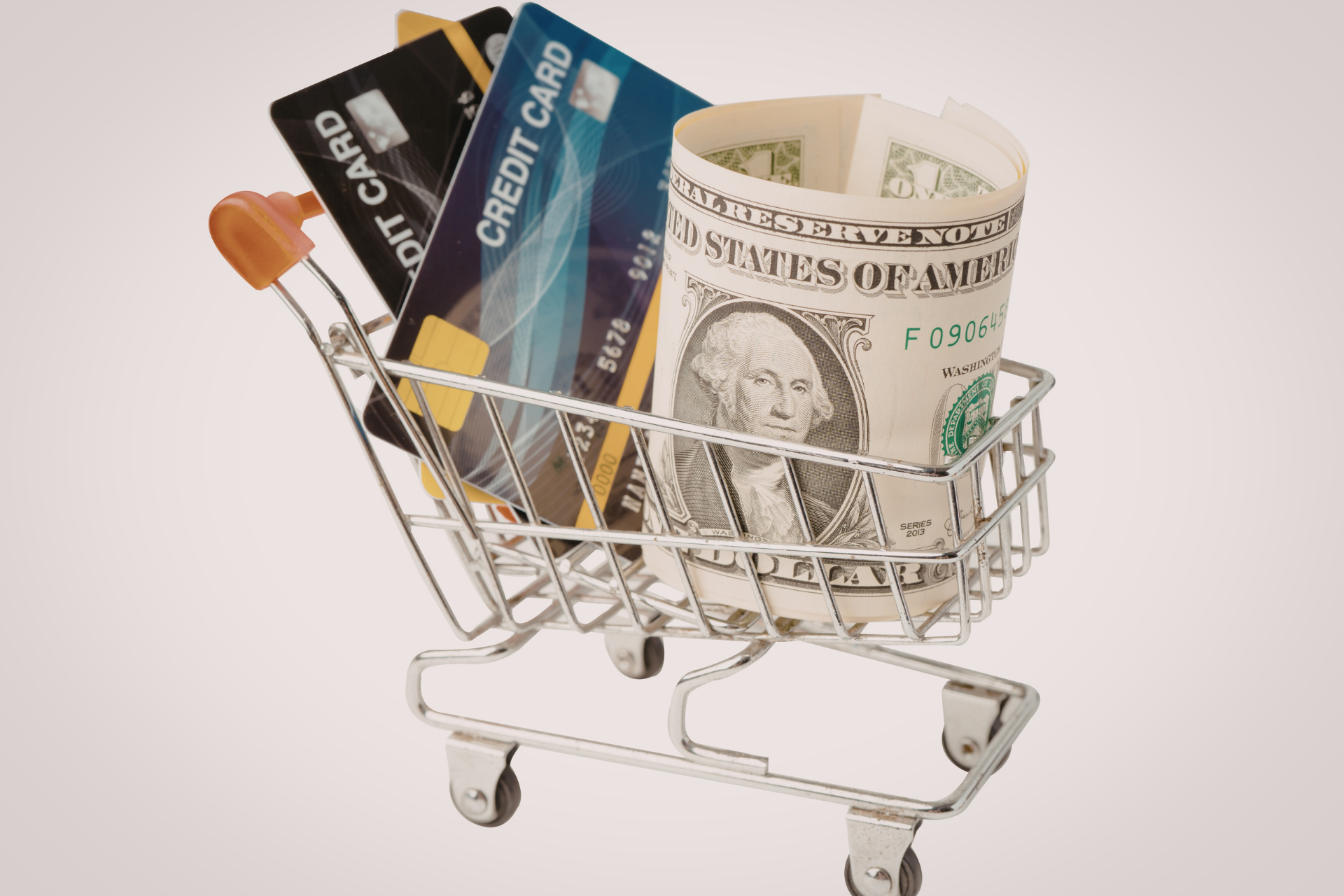 A small shopping cart containing two credit cards and a rolled-up US dollar bill.