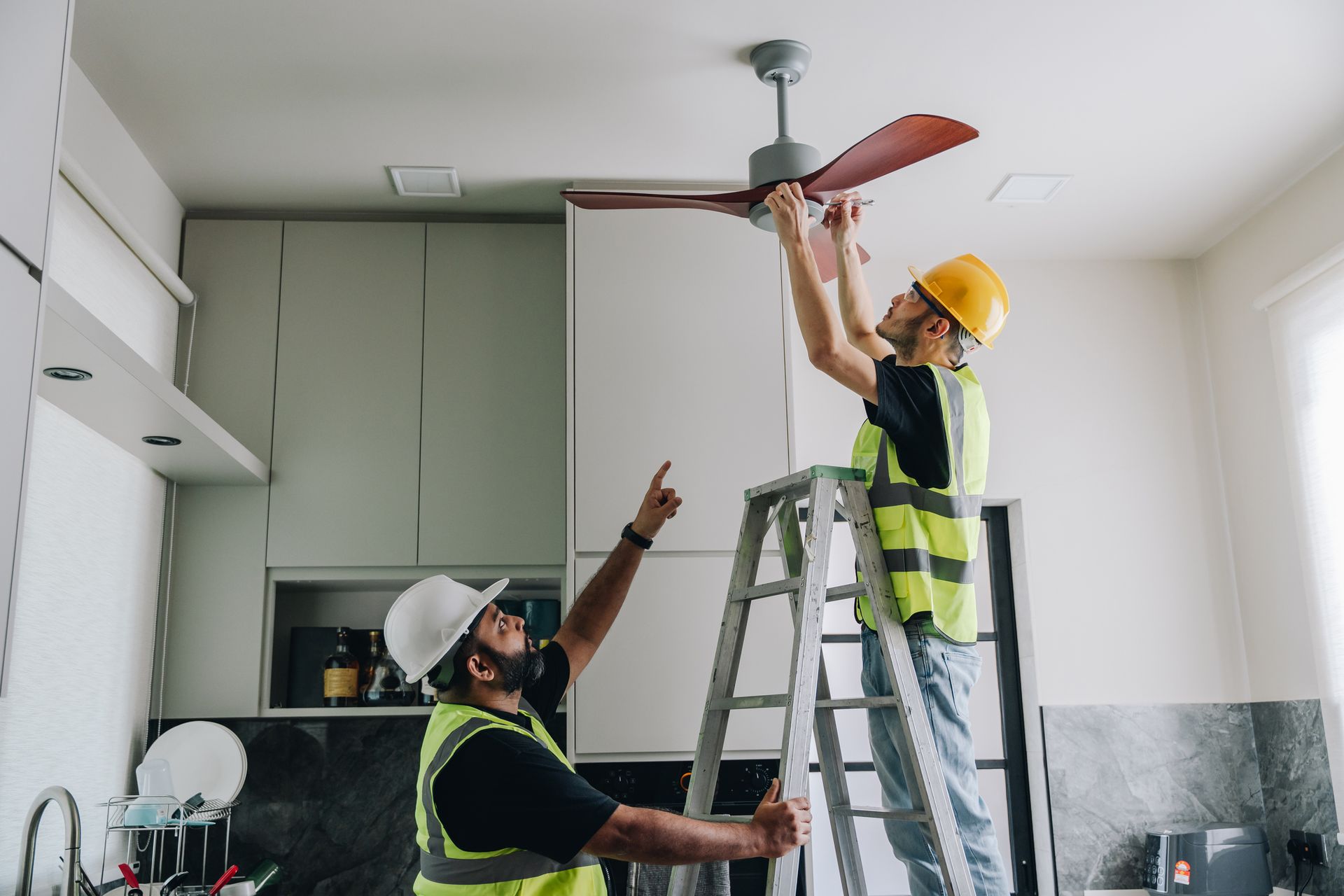 Two men are working on a ceiling fan in a kitchen.