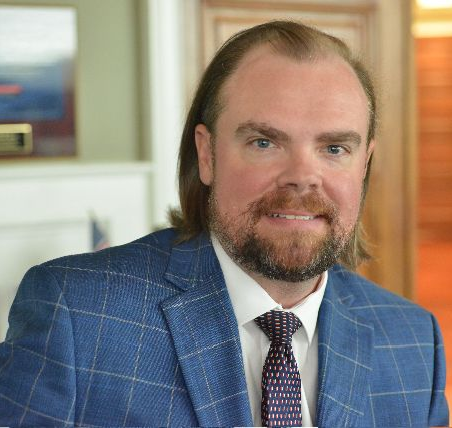 Man in blue plaid suit, smiling; indoors with painting and wooden door.
