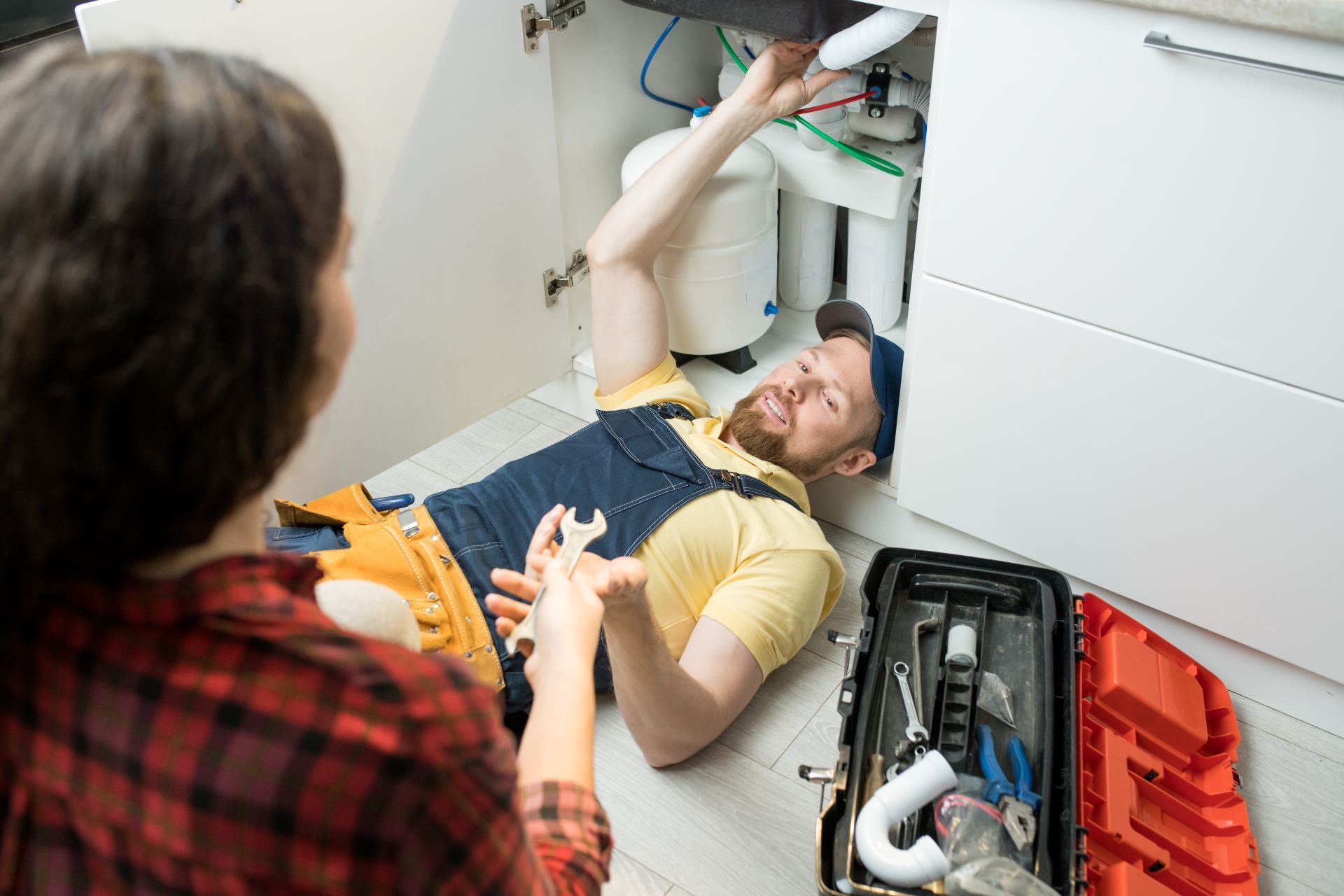 Plumber fixing pipes under kitchen sink while a person looks on.