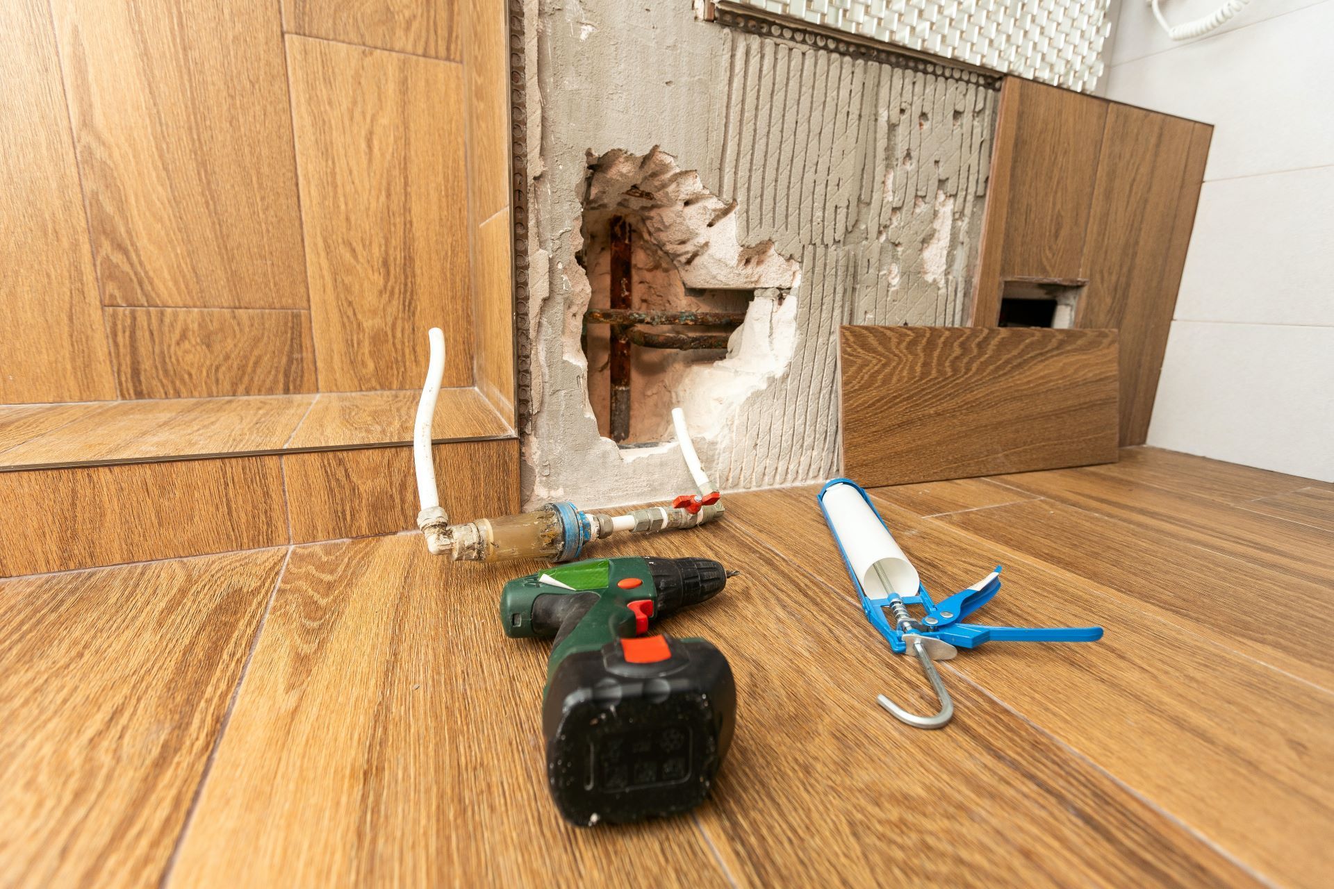 A power drill and caulk gun sit on a wood-look tiled floor in front of a hole in a tiled wall under construction.