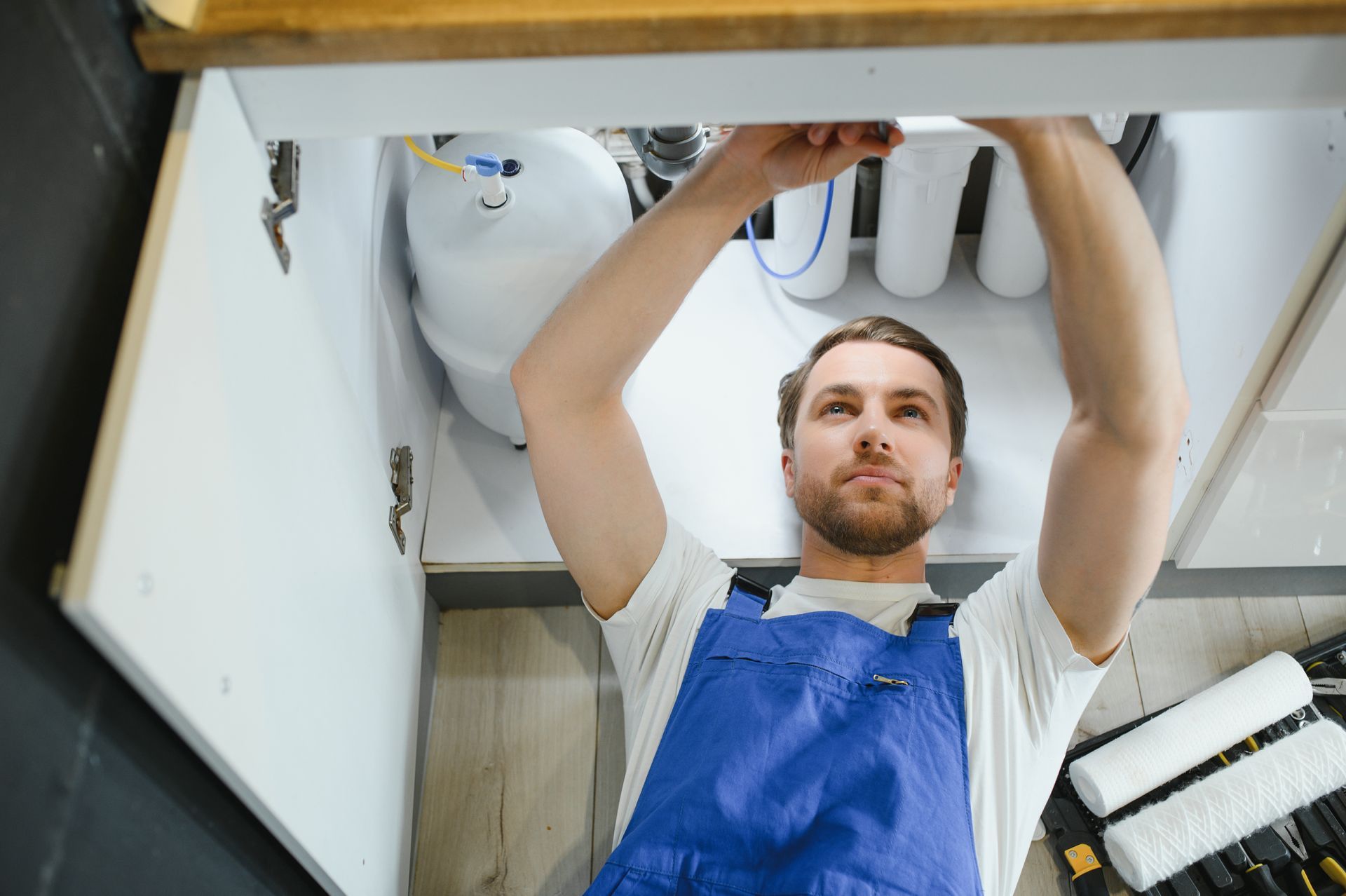 Plumber in blue overalls repairing pipes under a sink cabinet