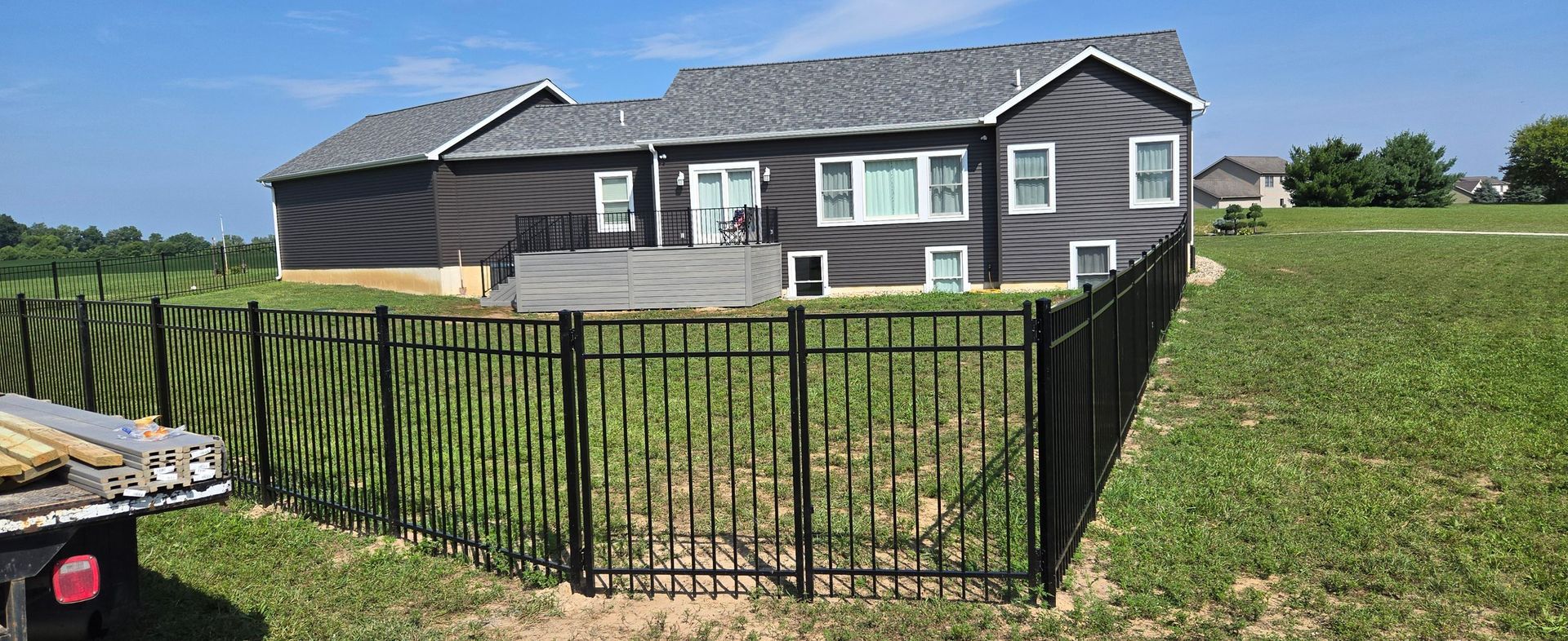 A dark-sided house with a back deck stands behind a black metal fence in a grassy yard on a sunny day.
