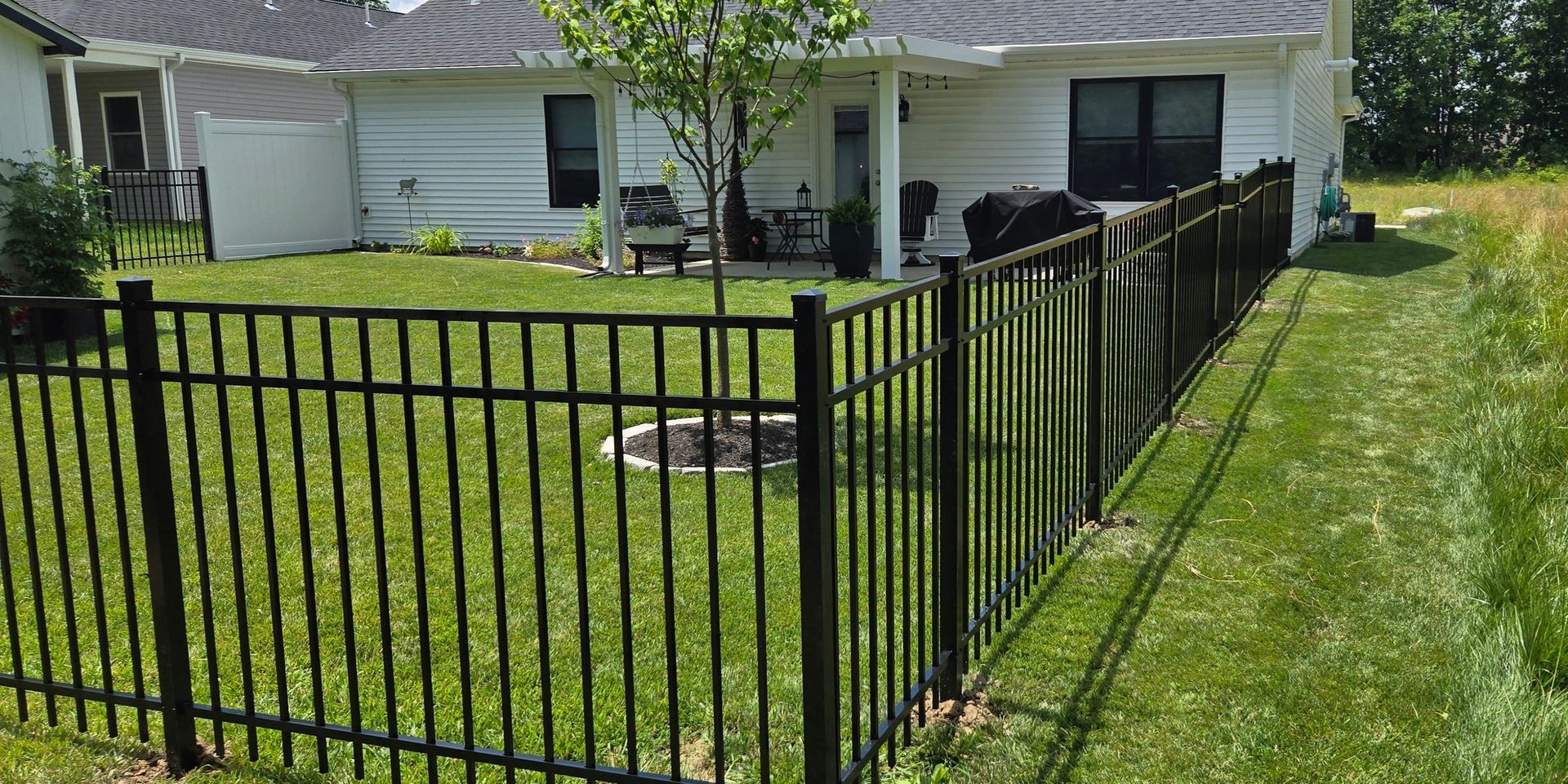 A black metal fence encloses a green grassy backyard with a tree and a white house with a patio in the background.