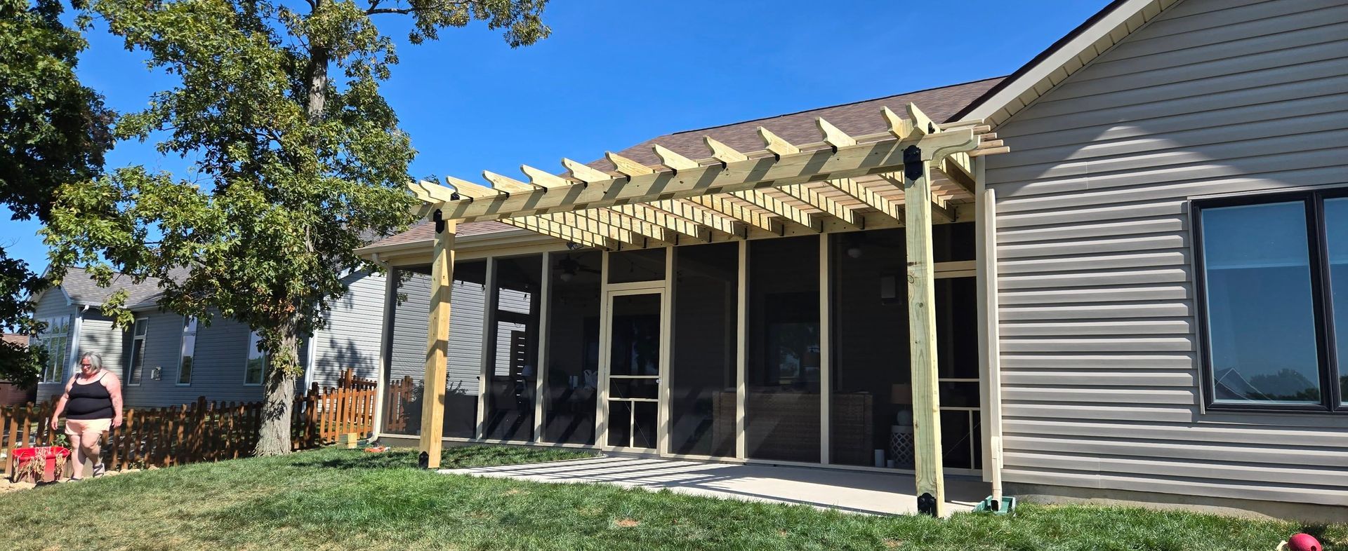 A beige house with a newly constructed wooden pergola over a screened-in porch, set in a sunny backyard with green grass.