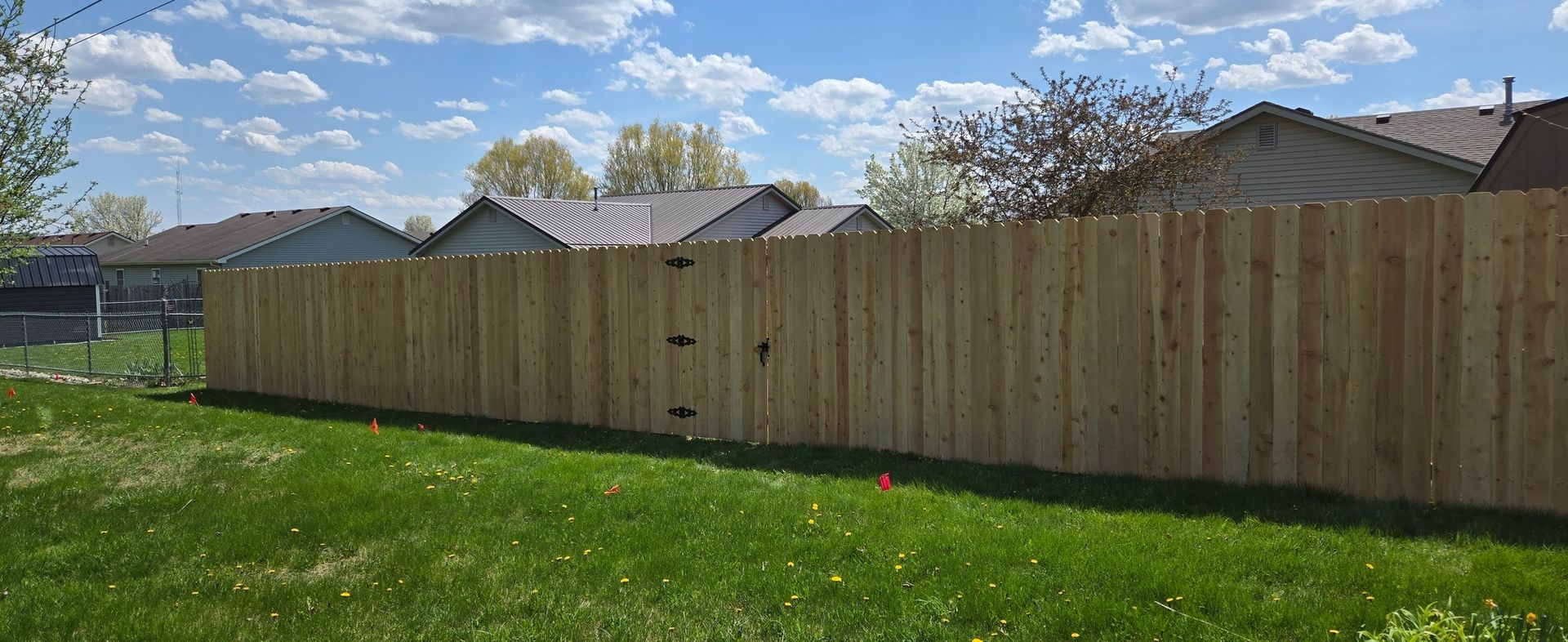 A new, vertical-plank wooden fence stands in a grassy backyard under a bright blue sky with scattered clouds.