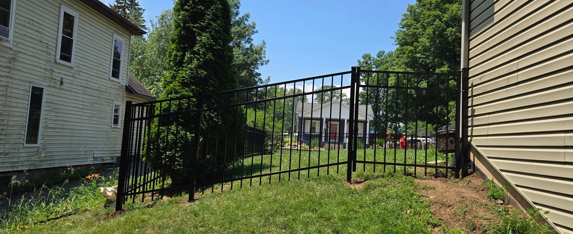 A black metal fence stretches between two light-colored houses on a grassy lawn under a sunny sky.