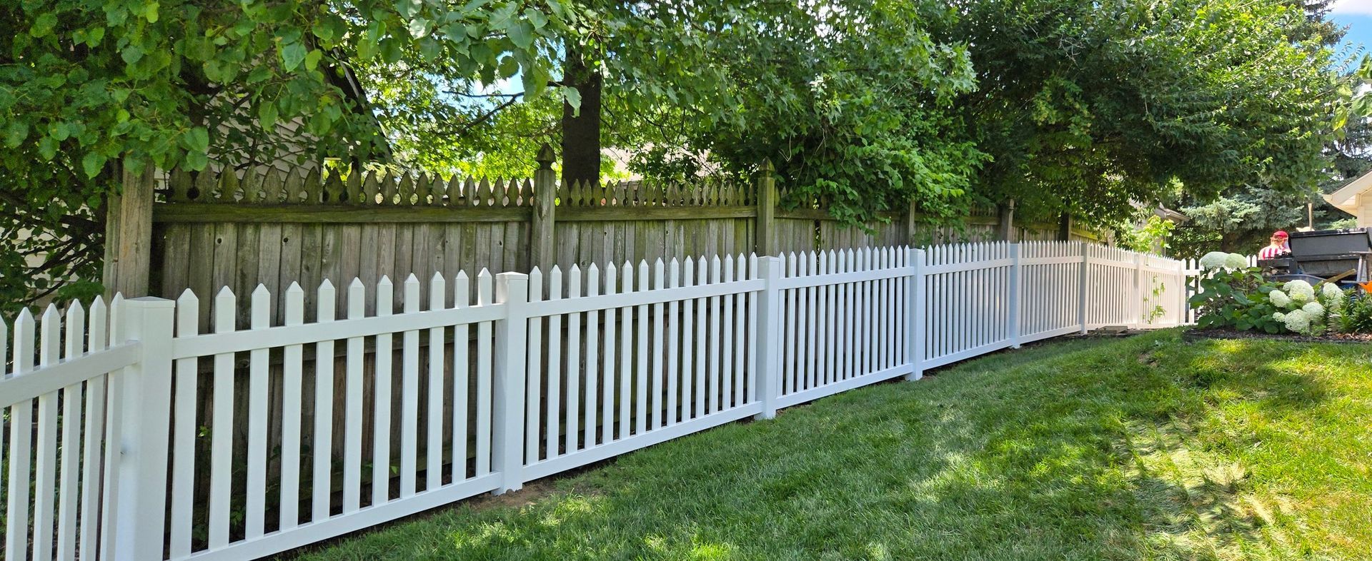A white vinyl picket fence stands in front of a wooden privacy fence, separated by green trees and a grassy lawn.
