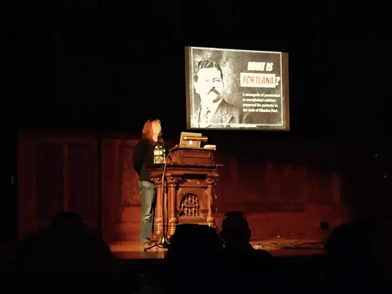 A woman stands at a podium in front of a large screen with a picture of a man on it.