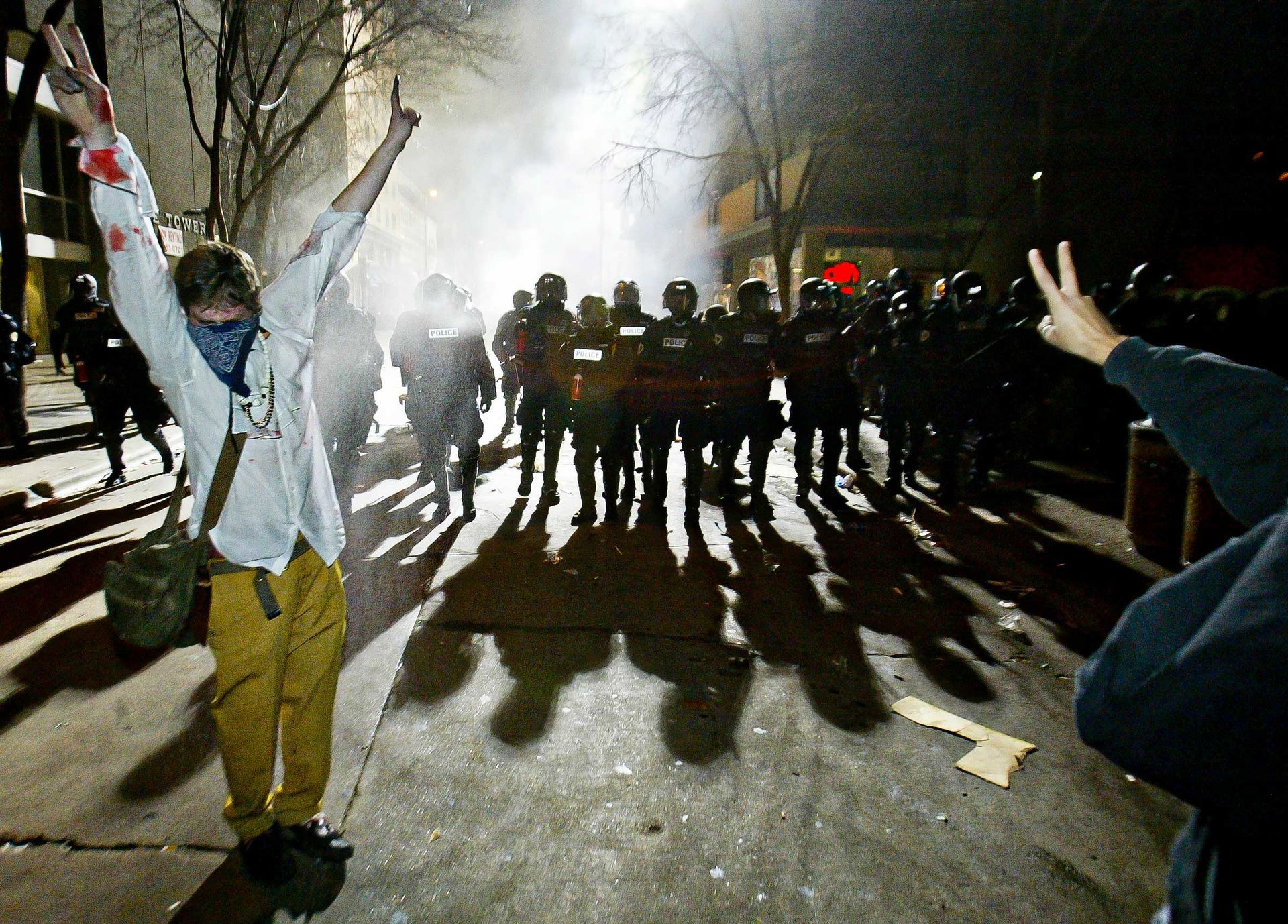 Protesters face a line of police in riot gear at night. One raises arms, another makes a peace sign.