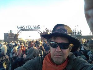A man wearing a hat and sunglasses is taking a selfie in front of a sign that says wasteland.
