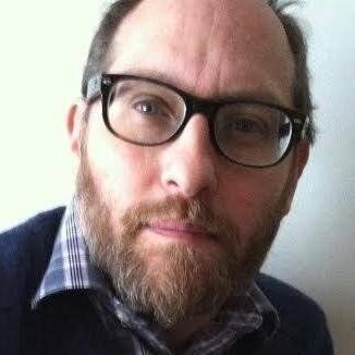 A man with a beard and glasses is looking at the camera.