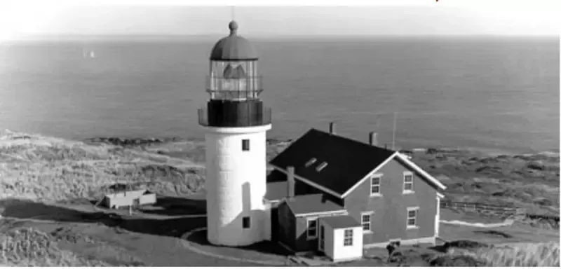 A black and white photo of a lighthouse and a house on a hill overlooking the ocean.