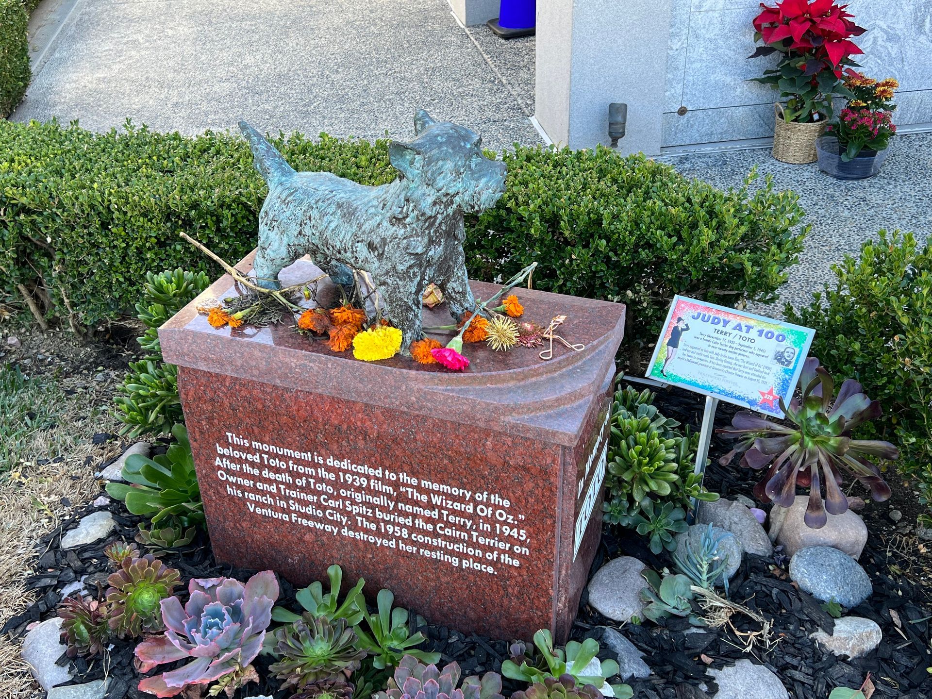 A statue of a dog is sitting on top of a red urn in a garden.