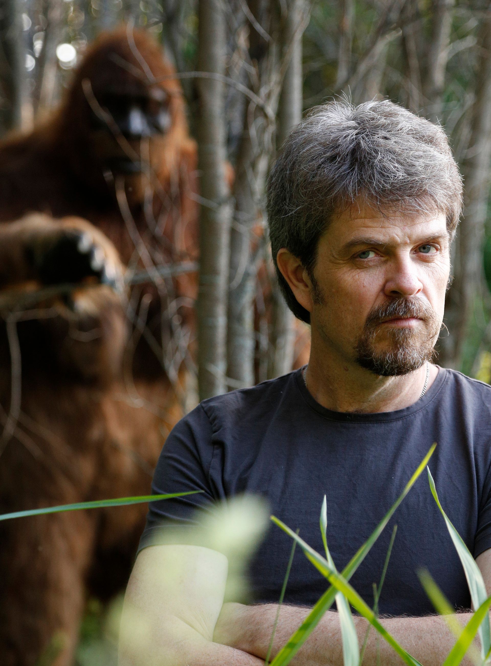 A man with a beard is standing in the woods with a stuffed animal in the background.