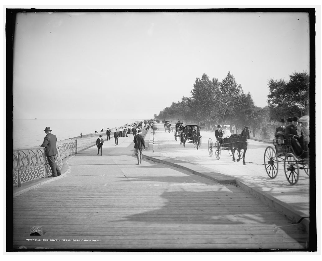 A black and white photo of a horse drawn carriage on a sidewalk.