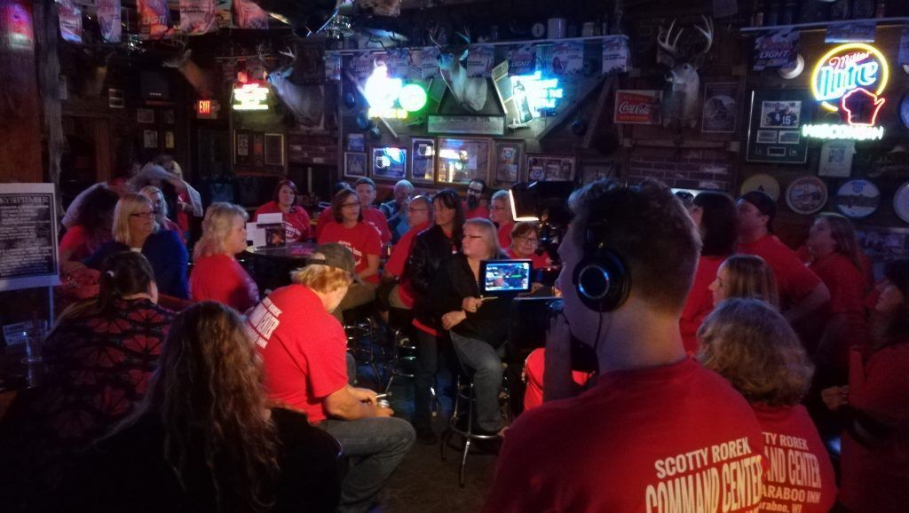 A man wearing headphones is standing in front of a crowd of people in a bar.