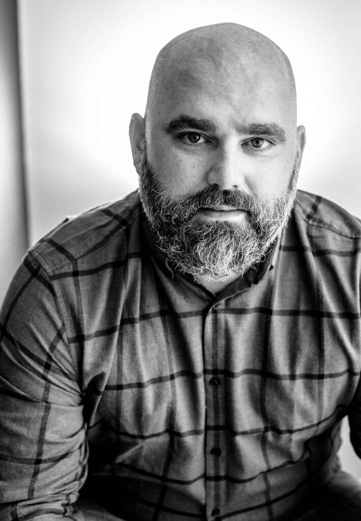 A black and white photo of a bald man with a beard wearing a plaid shirt.