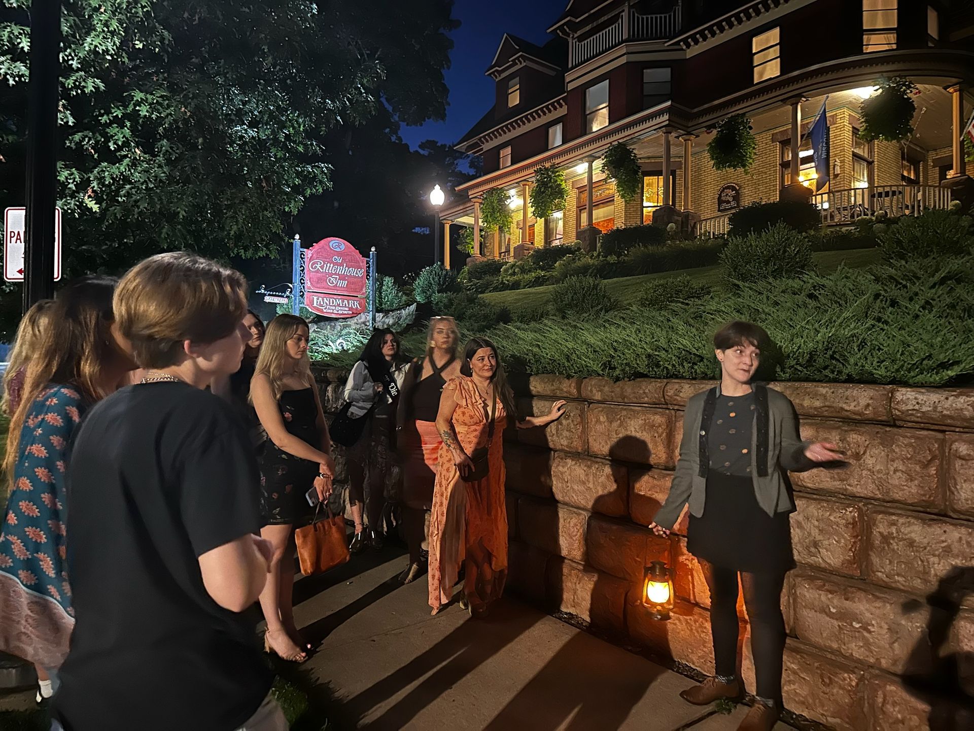 People on a nighttime walking tour, standing by a stone wall in front of a large lit Victorian building.