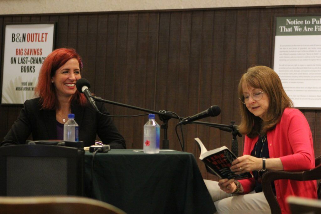 Two women are sitting at a table reading a book in front of microphones.