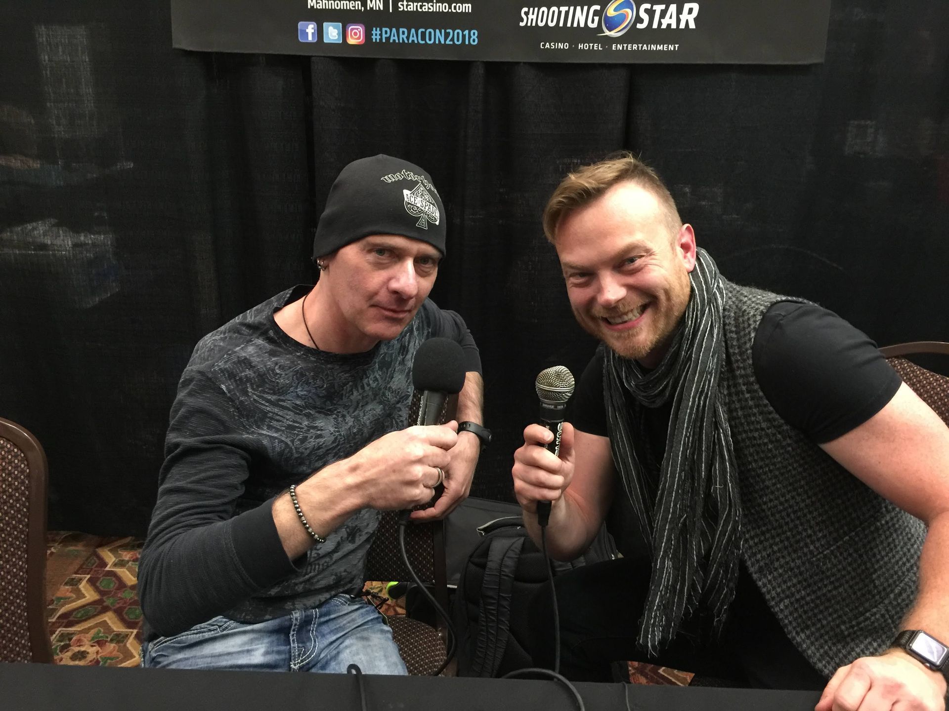 Two men are sitting at a table holding microphones and smiling.