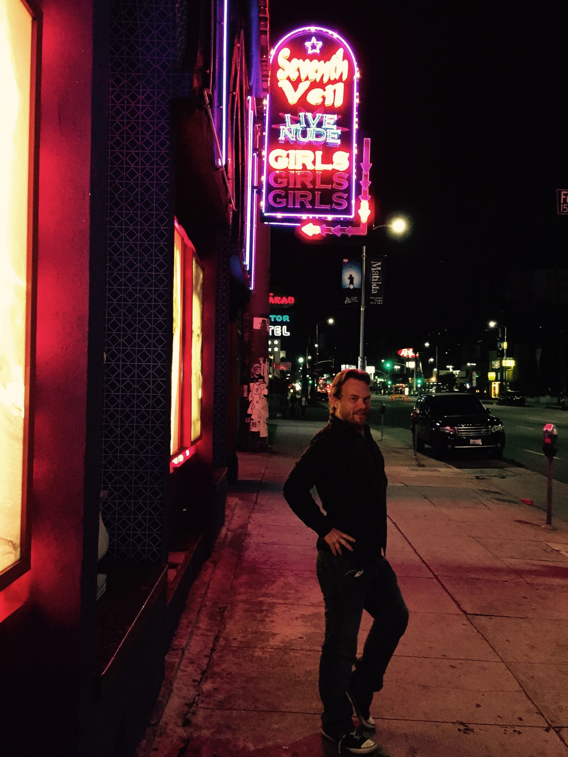 A man standing on a sidewalk in front of a neon sign that says girls