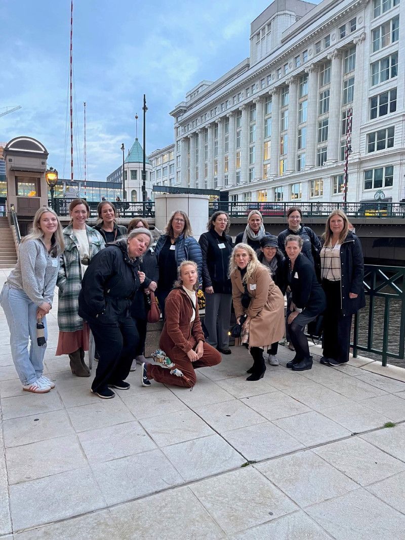 Group of people posing by a building and water. Some are crouching, smiling. Cloudy day.