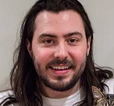 A man with long hair and a beard is holding a cat and smiling.