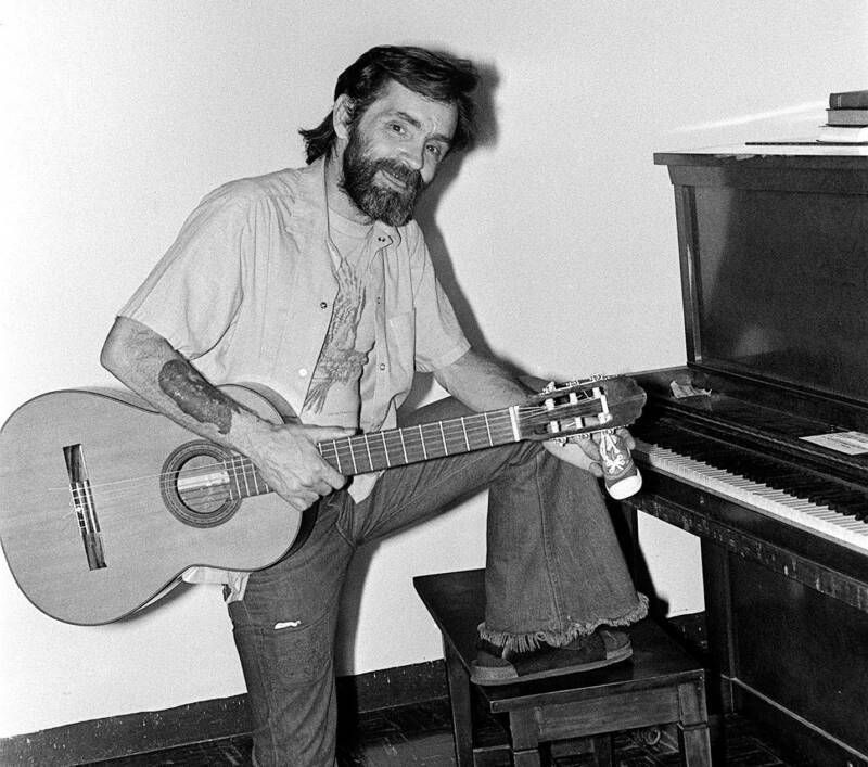 A man with a beard is holding a guitar next to a piano