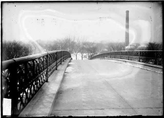 A black and white photo of a bridge with a chimney in the background