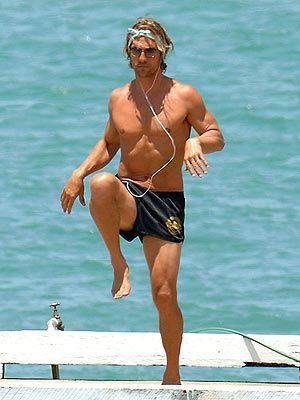 A shirtless man is doing exercises on a dock near the ocean.