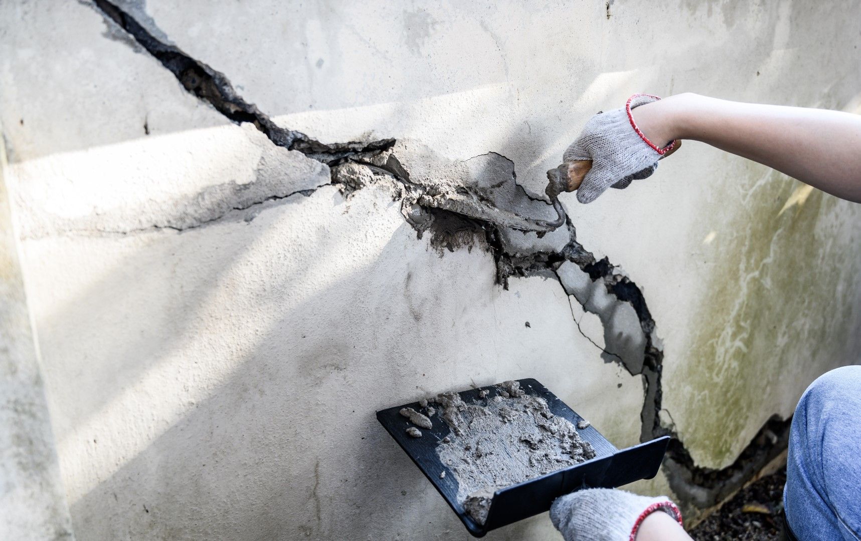 Person using a trowel to repair a large crack in a concrete wall, outdoors.