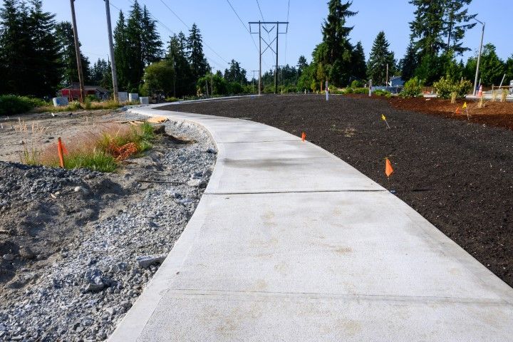 Concrete sidewalk curving through a construction zone, with dark soil and power lines.